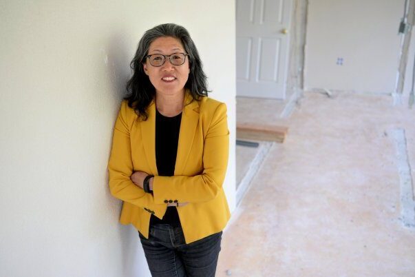 Susun Kim stands next to a wall in a yellow jacket and black blouse and pants. She has her arms folded and she is smiling