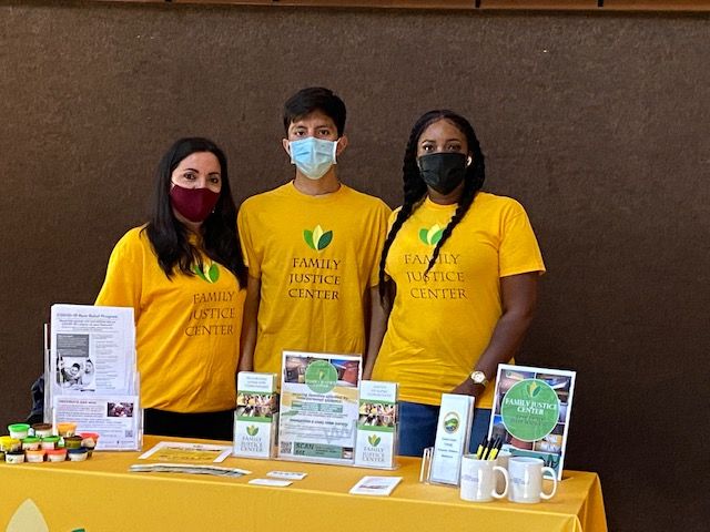 two young women and a young man, all in yellow Family Justice Center t-shirts, stand behind a table with pamphlets about safety and resources.