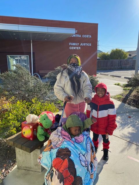 A woman stands in front of the family justice center holding a stroller with a toddler in it. She is wearing a bandanna over her mouth and nose but looks to be smiling. A child stands next to her smiling. All three are wearing warm clothes. There is a pile of new-looking toys on the low wall next to the stroller.