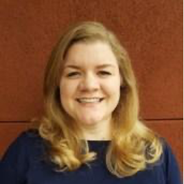 Natalie smiles at the camera. She has shoulder-length blond hair and wears a blue sweater.