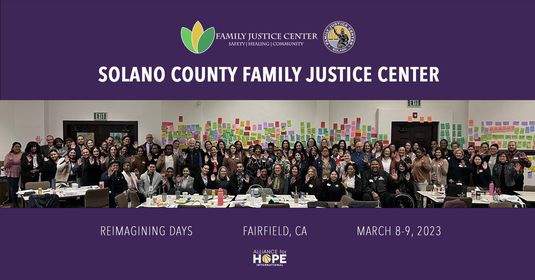 a large group of people, staff of Solano County nonprofit and government agencies, smile standing in front of a wall covered in post-it notes describing values and services of the ideal Family Justice Center