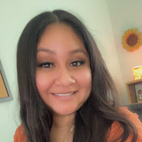 cropped-Mayra-Sanchez-1.jpg Myra smiles at the camera. She wears a black blouse and orange cardigan sweater.