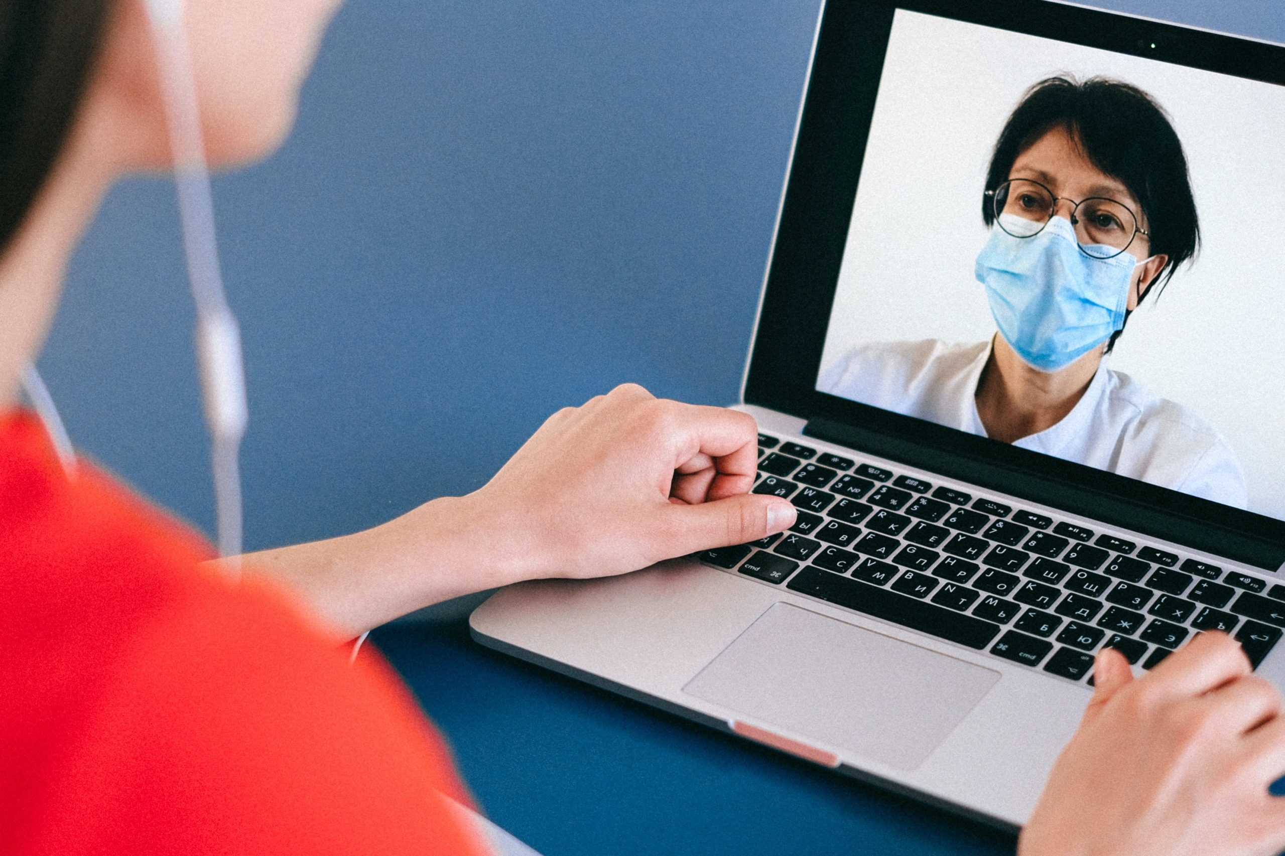 A person in an orange shirt and earphones faces a laptop screen showing a medical professional in white coat and blue surgical mask.