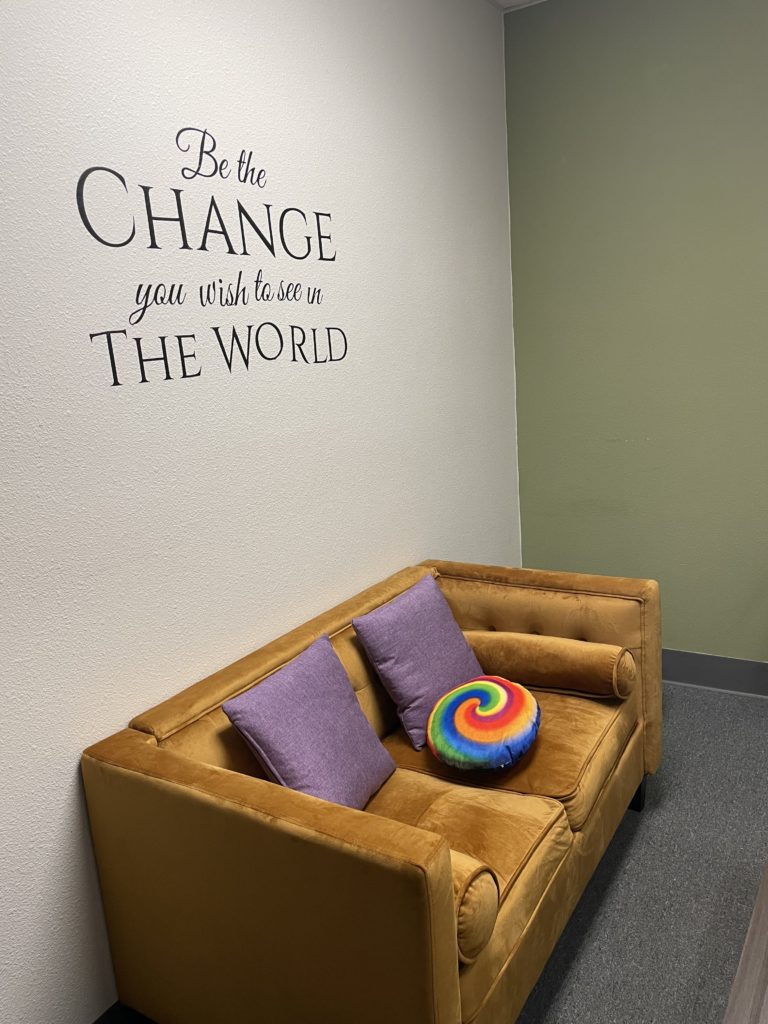 Gold couch with rainbow pillow under wall sign reading "Be the Change you want to see in the World"