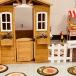 The play house front has two windows, a half door, white picket fence and mailbox. Nearby a farmers market stand invites kids to grocery shop.
