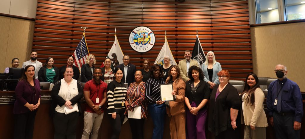 22 Family Justice Center partners stand in meeting room in front of the Solano County seal. Director Reina Beverly-Sandoval holds the Solano Board of Supervisors DVAM Proclamation.