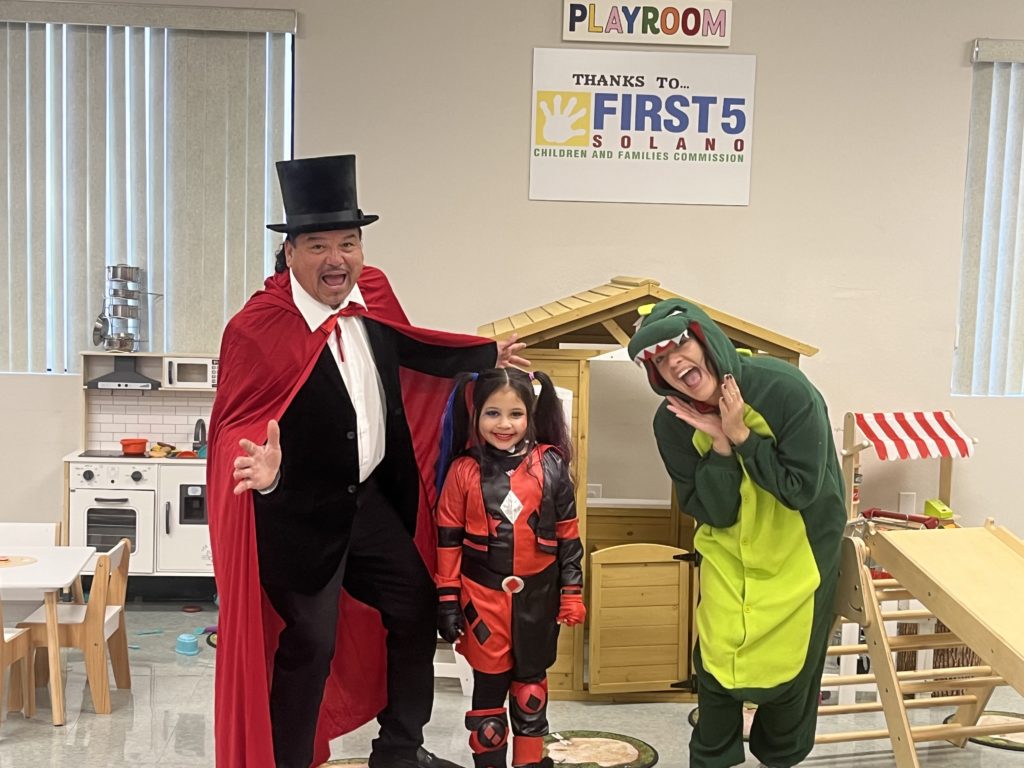 A man in a top hat and red cape, little girl in a red and black suit, and woman in Barney costume strike poses in the playroom.