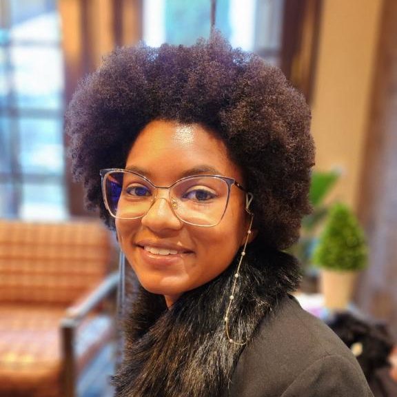 Cyrina wears clear-framed glasses and has brown curly hair and a fleece-collared jacket. She smiles at the camera.