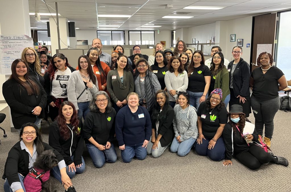 34 staff (30 women, 4 men) from the 4-center network of Family Justice Centers are grouped in a conference room smiling at the camera. Many of them wear black sweatshirts or t-shirts with the FJC lotus logo. In the front row, eight people kneel; the rest stand in rows behind them. A service dog sits in front of a woman at left.